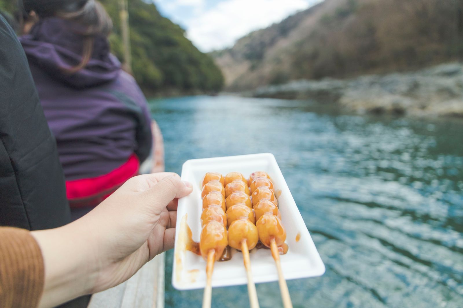 Bàn tay cầm đĩa dango mitarashi bên dòng sông xanh ở Arashiyama, Kyoto