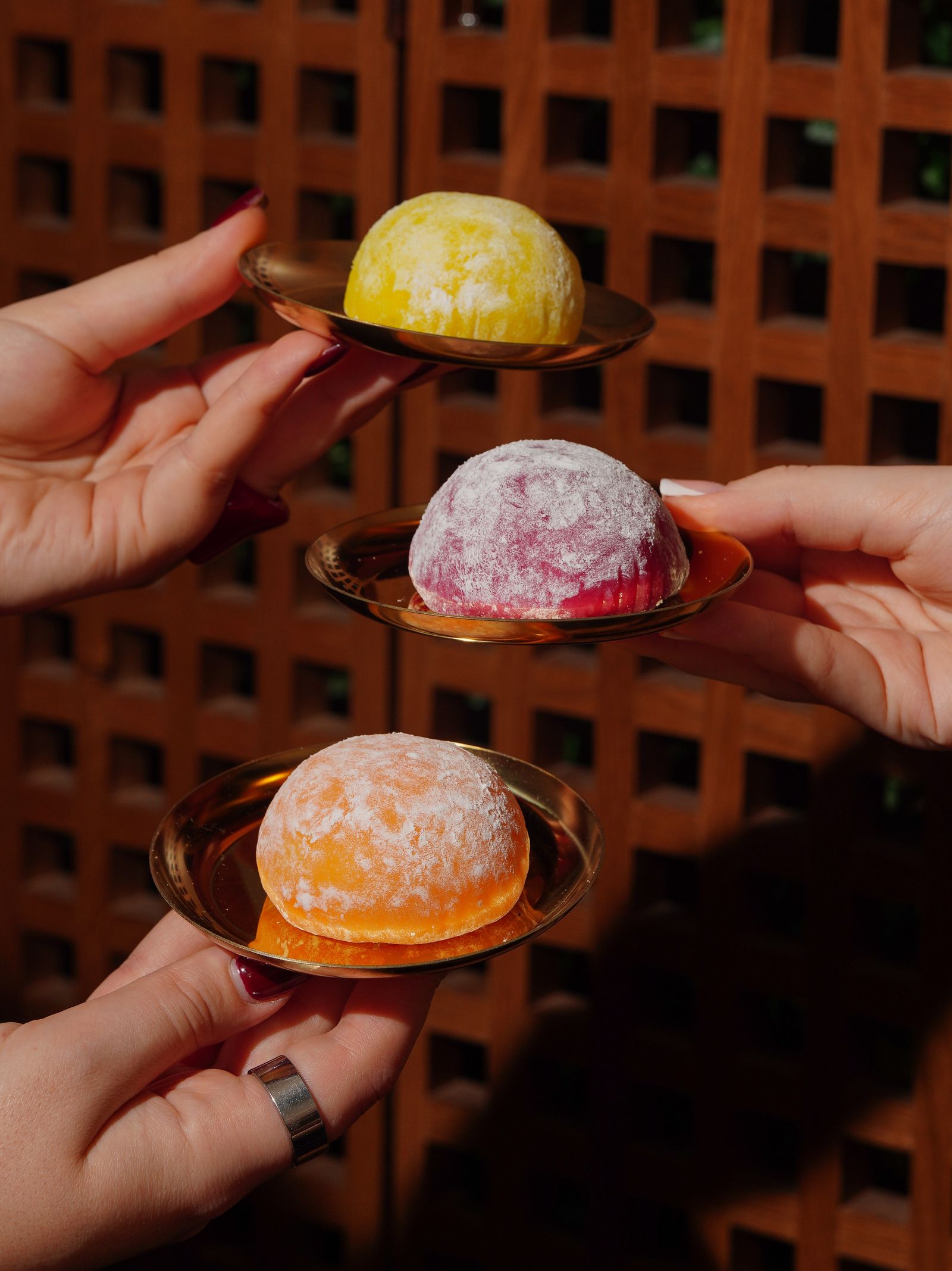 Ba bàn tay giơ lên ba chiếc mochi vàng, hồng, cam trên đĩa gỗ trước hàng rào gỗ đỏ kiểu Nhật